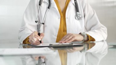Beyaz bir sağlık önlüğünün altına sarı bluz giyen kadın doktor, bir pano kullanıyor ve yakın plandaki cam masaya notlar alıyor. Tıp konsepti
