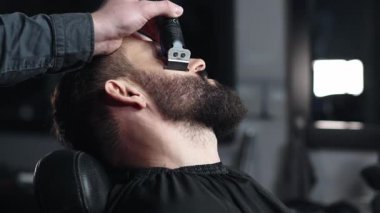 Profesyonel berber erkek müşterinin sakalını elektrikli budayıcı kullanarak dikkatlice şekillendiriyor, modern berber dükkanında titiz bir bakım ve stil sağlıyor.