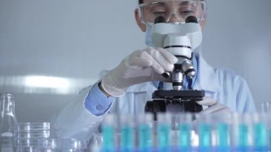 Laboratuvar önlüğü, maskesi, beyaz eldivenleri ve güvenlik gözlüğü takan bilim adamı bir kadın laboratuarda test tüpleriyle çevrili bir mikroskop kullanıyor. Portre görüntüsü. Tıp ve bilim konsepti.