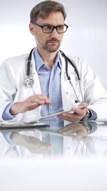 Beyaz önlük, gözlük ve steteskop altında mavi gömlekli doktor modern sağlık ortamında dijital tablet kullanıyor. Hasta verileriyle, dikey portre görüntüsüyle etkileşimde. Tıp ve Bilim.