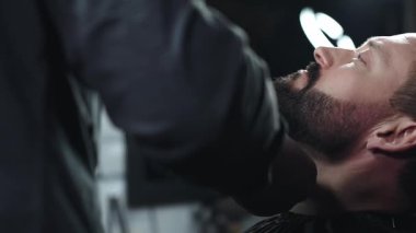 Profesyonel berber, modern berber dükkanında yüksek kaliteli tımar hizmetleri sunarak gevşetilmiş müşterinin sakalını elektrikli jiletle dikkatlice buduyor.