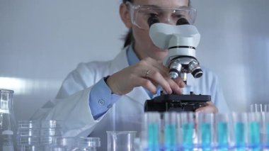 Laboratuvar önlüğü ve güvenlik gözlüğü takan bilim adamı bir kadın laboratuarda test tüpleriyle çevrili bir mikroskop kullanıyor. Tıp ve bilim konsepti.