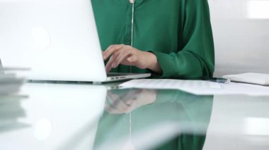 Yeşil günlük bluz giyen iş kadını dizüstü bilgisayarda daktilo kullanıyor, vergileri ve teftiş raporlarını hazırlıyor ve cam ofis masasında belgeler bulunuyor. Vergiler, iş denetimleri.
