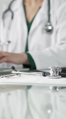 Stetoskop ve ilaç geçmişi kayıtları cam bir masanın üzerinde duruyor. Klinik ofis arka planında dizüstü bilgisayarla çalışan bir doktorun önünde dikey yakın plan görüntüsü. Tıp konsepti.