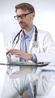 Doktor bey, steteskop, mavi gömlek ve laboratuvar önlüğü giyen doktor klinikte dizüstü bilgisayarlı cam bir masa ve dikey portre manzaralı el kol hareketi yapıyor. Tıp ve Bilim.