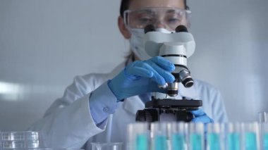 Laboratuvar önlüğü, maske, mavi eldiven ve güvenlik gözlüğü takan bilim adamı kadın, laboratuarda test tüpleriyle çevrili bir mikroskobu ayarlıyor. Tıp ve bilim konsepti.