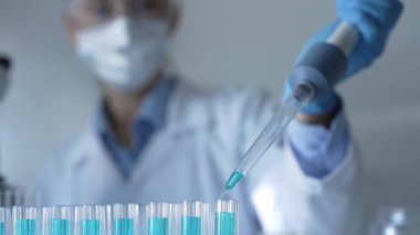 Laboratuvar önlüğü, maskesi ve güvenlik gözlüğü takan kadın araştırmacı test tüpüne mavi bir sıvı atmak için mikropipet kullanıyor. Tıp ve bilim konsepti.