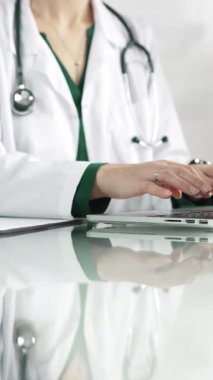 Doktor önlüğünün altında yeşil bluz giyen bir kadın doktor sağlık hizmetlerinde teknoloji kullanıyor. Klinikteki cam bir masanın üstünde dizüstü bilgisayara dikey bir bakış açısı olan ellerin yakın çekimi. Tıbbi konsept.