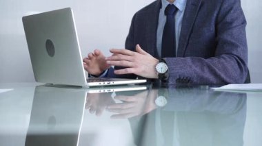 Mavi ceket giyen işadamı, modern bir ofisteki cam bir masadaki dizüstü bilgisayarla çevrimiçi iletişime odaklanarak üretkenlik ve konsantrasyon gösteriyor. İş veya denetim kavramı.
