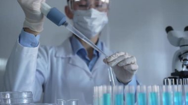 Laboratuvar önlüğü, maske ve güvenlik gözlüğü takan bilim adamı kadın araştırmacı test tüplerine sıvı atmak için bir mikropipet kullanıyor. Tıp ve bilim konsepti.