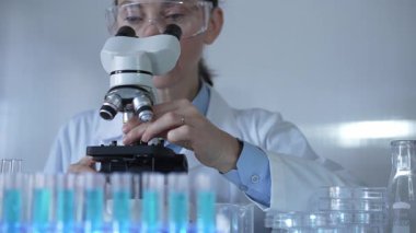 Laboratuvar önlüğü ve güvenlik gözlüğü takan bilim adamı bir kadın laboratuarda test tüpleriyle çevrili bir mikroskop kullanıyor. Tıp ve bilim konsepti.