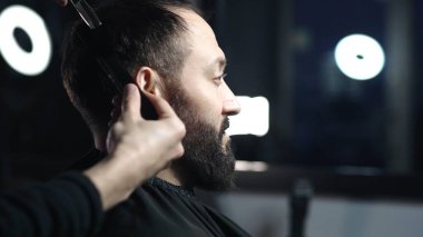 Profesyonel berber, modaya uygun bir berber dükkanında profesyonel bir makasla, sakallı bir adam için modern bir saç stili hazırlayan bir müşterinin saçını ustaca kesiyor.