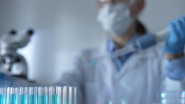 Mavi sıvıyla test tüpleri laboratuvar ortamında, yakın görüntüde. Kadın araştırmacı, arka planda bir mikro pipet kullanıyor. Tıp ve Bilim.