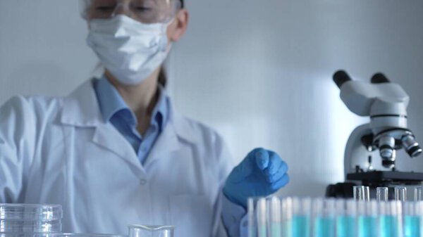Scientist woman, wearing a lab coat, mask, blue gloves and safety glasses, is using a micropipette to drop a liquid into a test tubes. Medicine and science.