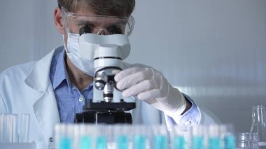 Laboratuvar önlüğü, maskesi, beyaz eldivenleri ve güvenlik gözlüğü takan erkek bilim adamı laboratuarda test tüpleriyle çevrili bir dürbün kullanıyor..