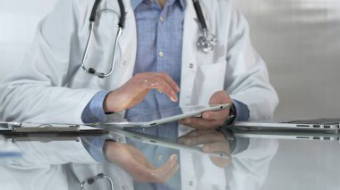 Tıbbi beyaz önlüğün altına mavi gömlek giyen doktor, modern sağlık ortamında dijital tablet kullanıyor, hasta verileriyle etkileşimde, yakın plan. İlaç ve sağlık hizmetleri.
