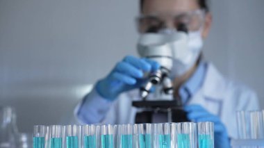 Mavi bir sıvıyla dolu laboratuvar tüpleri odaklanmış, yakından bakıyor. Kadın araştırmacı, arka planda mikroskop kullanıyor. Tıp ve bilim konsepti.