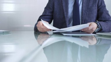 Mavi ceketli bir işadamı modern ofis ortamında cam bir masada belgeleri tutarken önemli evrakları inceliyor. İş adamları veya denetim kavramı.
