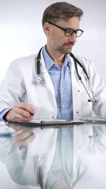 Mavi gömlekli doktor, tıp önlüğünün altında bir pano kullanıyor ve bir tıp bürosundaki cam masaya, dikey portre görüntüsüne notlar alıyor. Tıp ve Bilim.