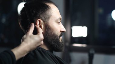 Profesyonel berber, modaya uygun bir berber dükkanında profesyonel bir makasla, sakallı bir adam için modern bir saç stili hazırlayan bir müşterinin saçını ustaca kesiyor.