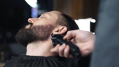 Profesyonel berber erkek müşterinin sakalını elektrikli budayıcı kullanarak dikkatlice şekillendiriyor, modern berber dükkanında titiz bir bakım ve stil sağlıyor.