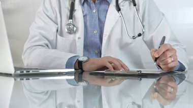 Beyaz bir sağlık önlüğünün altına mavi gömlek giyen erkek doktor, bir not panosu kullanıyor ve yakın plandaki cam masaya notlar alıyor. Tıp konsepti.