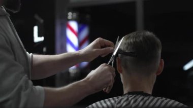 Profesyonel berber erkek müşterinin saçını şık bir berber dükkanında makas ve tarak kullanarak ustaca kesiyor ve arka planda belirgin bir şekilde berber direği sergiliyor.