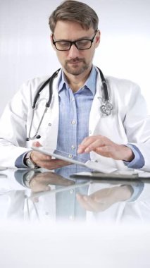 Beyaz önlük, gözlük ve steteskop altında mavi gömlekli doktor modern sağlık ortamında dijital tablet kullanıyor. Hasta verileriyle, dikey portre görüntüsüyle etkileşimde. Tıp ve Bilim.