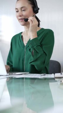 Yeşil bluzlu iş kadını, modern ofis ortamında, dikey portrede dizüstü bilgisayarla etkin bir şekilde çalışırken, bir kulaklık takıyor. İş adamları, çağrı merkezi kavramı
