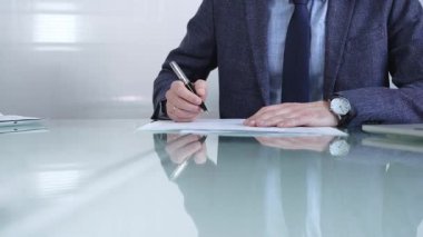 Çağdaş bir ofis ortamında modern cam bir masanın üzerinde profesyonel giyimli bir işadamı kalemle kontrat imzalıyor. İş adamları veya denetim kavramı