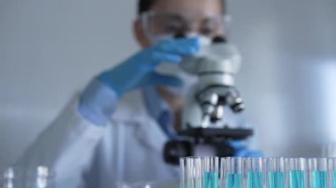 Laboratuvar önlüğü, maske, mavi eldiven ve güvenlik gözlüğü takan bilim adamı kadın, laboratuarda test tüpleriyle çevrili bir mikroskobu ayarlıyor. Tıp ve bilim konsepti.