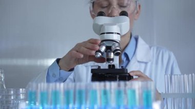Laboratuvar önlüğü ve güvenlik gözlüğü takan bilim adamı bir kadın laboratuarda test tüpleriyle çevrili bir mikroskop kullanıyor. Tıp ve bilim konsepti.