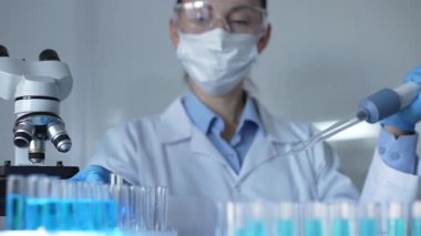 Laboratuvar önlüğü, maske ve güvenlik gözlüğü takan bilim adamı kadın araştırmacı test tüplerine sıvı atmak için bir mikropipet kullanıyor. Tıp ve bilim konsepti.