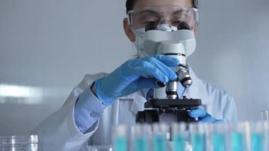 Laboratuvar önlüğü, maske, mavi eldiven ve güvenlik gözlüğü takan bilim adamı kadın, laboratuarda test tüpleriyle çevrili bir mikroskobu ayarlıyor. Tıp ve bilim konsepti.