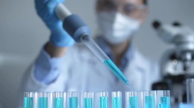 Laboratuvar önlüğü, maskesi ve güvenlik gözlüğü takan kadın araştırmacı test tüpüne mavi bir sıvı atmak için mikropipet kullanıyor. Tıp ve bilim konsepti.