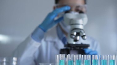 Mavi bir sıvıyla dolu laboratuvar tüpleri odaklanmış, yakından bakıyor. Kadın araştırmacı, arka planda mikroskop kullanıyor. Tıp ve bilim konsepti.