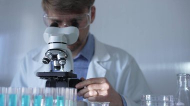 Laboratuvar önlüğü, maskesi ve güvenlik gözlüğü takan bir bilim adamı laboratuvar ortamında test tüpleriyle çevrili bir mikroskop kullanıyor. Tıp ve bilim konsepti.