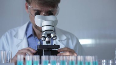 Laboratuvar önlüğü, maskesi ve güvenlik gözlüğü takan bir bilim adamı laboratuvar ortamında test tüpleriyle çevrili bir mikroskop kullanıyor. Tıp ve bilim konsepti.