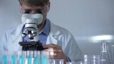 Laboratuvar önlüğü, maskesi ve güvenlik gözlüğü takan bir bilim adamı laboratuvar ortamında test tüpleriyle çevrili bir mikroskop kullanıyor. Tıp ve bilim konsepti.