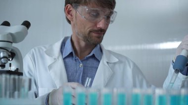 Laboratuvar önlüğü ve güvenlik gözlüğü takan bilim adamı deney tüpüne sıvı atmak için mikropipet kullanıyor. Portre görüntüsü. Tıp ve Bilim.