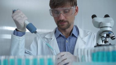 Laboratuvar önlüğü ve güvenlik gözlüğü takan bilim adamı deney tüpüne sıvı atmak için mikropipet kullanıyor. Portre görüntüsü. Tıp ve Bilim.