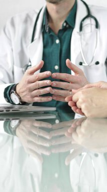 Steteskop ve laboratuvar önlüğü giyen doktor, masadaki bir hastaya dizüstü bilgisayarla teşhis koymayı açıklarken elleriyle işaret ediyor. Tıp konsepti.