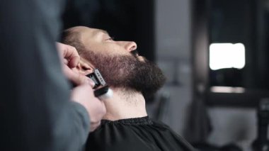 Profesyonel berber erkek müşterinin sakalını elektrikli budayıcı kullanarak dikkatlice şekillendiriyor, modern berber dükkanında titiz bir bakım ve stil sağlıyor.