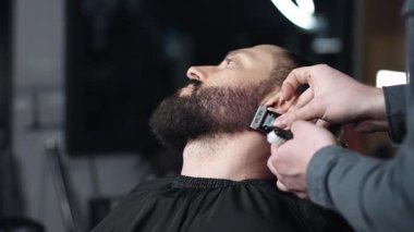 Profesyonel berber erkek müşterinin sakalını elektrikli budayıcı kullanarak dikkatlice şekillendiriyor, modern berber dükkanında titiz bir bakım ve stil sağlıyor.