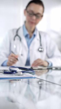 Stetoskop ve ilaç geçmişi kayıtları cam bir masanın üzerinde duruyor. Klinik ofis arka planında dizüstü bilgisayarla çalışan bir doktorun önünde dikey yakın plan görüntüsü. Tıp ve Bilim.
