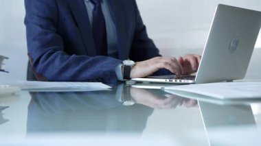 Mavi takım elbiseli iş adamı cam masada dizüstü bilgisayarda yazarken parlak bir ofiste oturuyor, üretkenlik ve modern çalışma ortamını gösteriyor. İş insanları kavramı.