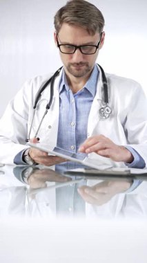 Beyaz önlük, gözlük ve steteskop altında mavi gömlekli doktor modern sağlık ortamında dijital tablet kullanıyor. Hasta verileriyle, dikey portre görüntüsüyle etkileşimde. Tıp ve Bilim.