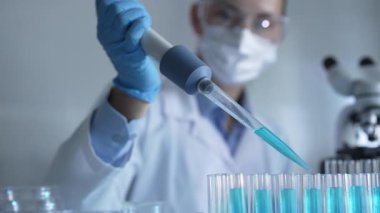 Laboratuvar önlüğü, maskesi ve güvenlik gözlüğü takan kadın araştırmacı test tüpüne mavi bir sıvı atmak için mikropipet kullanıyor. Tıp ve bilim konsepti.