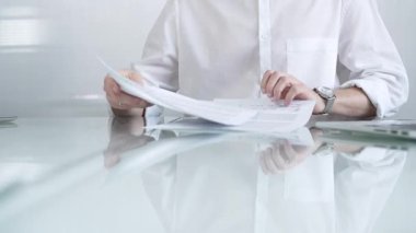 Beyaz gömlekli bir iş adamı, güncel ofis ortamında cam bir masada evrakları tutarken evrak işleriyle uğraşıyor. İş adamları konsepti.