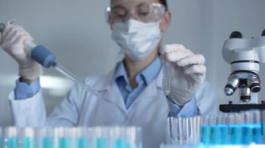 Laboratuvar önlüğü, maske ve güvenlik gözlüğü takan bilim adamı kadın araştırmacı test tüplerine sıvı atmak için bir mikropipet kullanıyor. Tıp ve bilim konsepti.